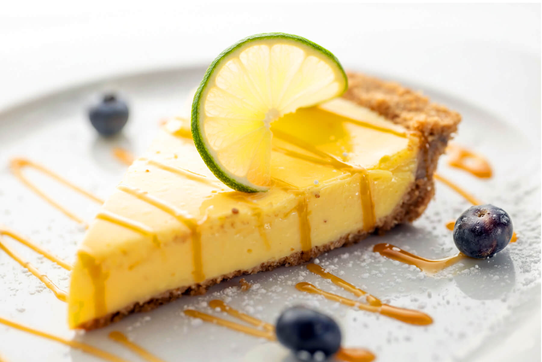 Slice of key lime pie with a lime wheel on top, drizzled with caramel sauce, and garnished with blueberries on a white plate.