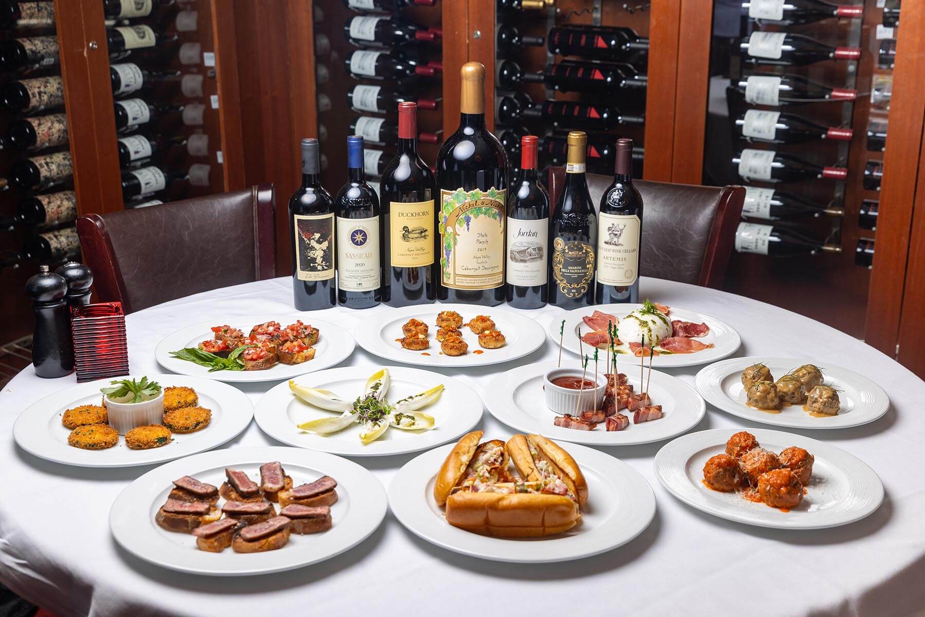 Table with assortment of appetizers and eight bottles of red wine in front of a wine rack in a dining room.