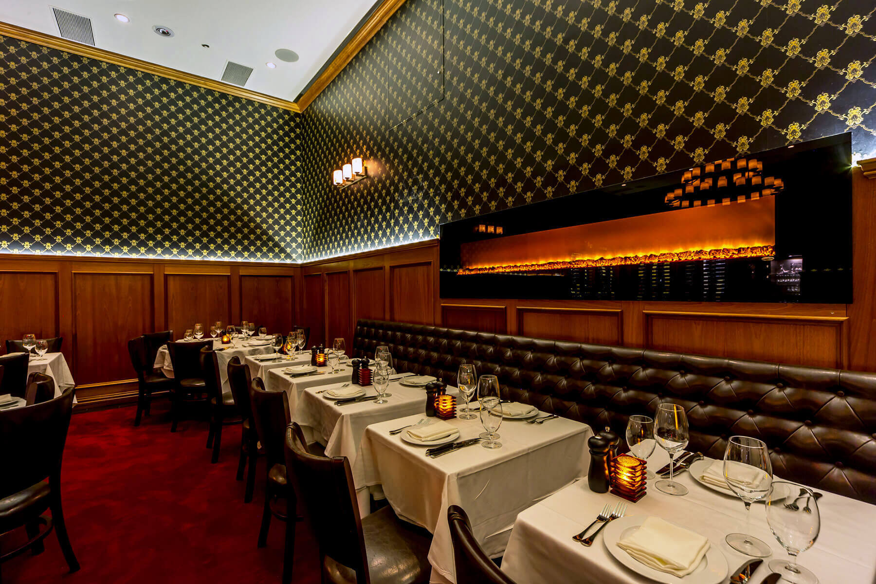 Elegant restaurant dining area with white tablecloths, leather chairs, patterned wallpaper, and a long modern fireplace.